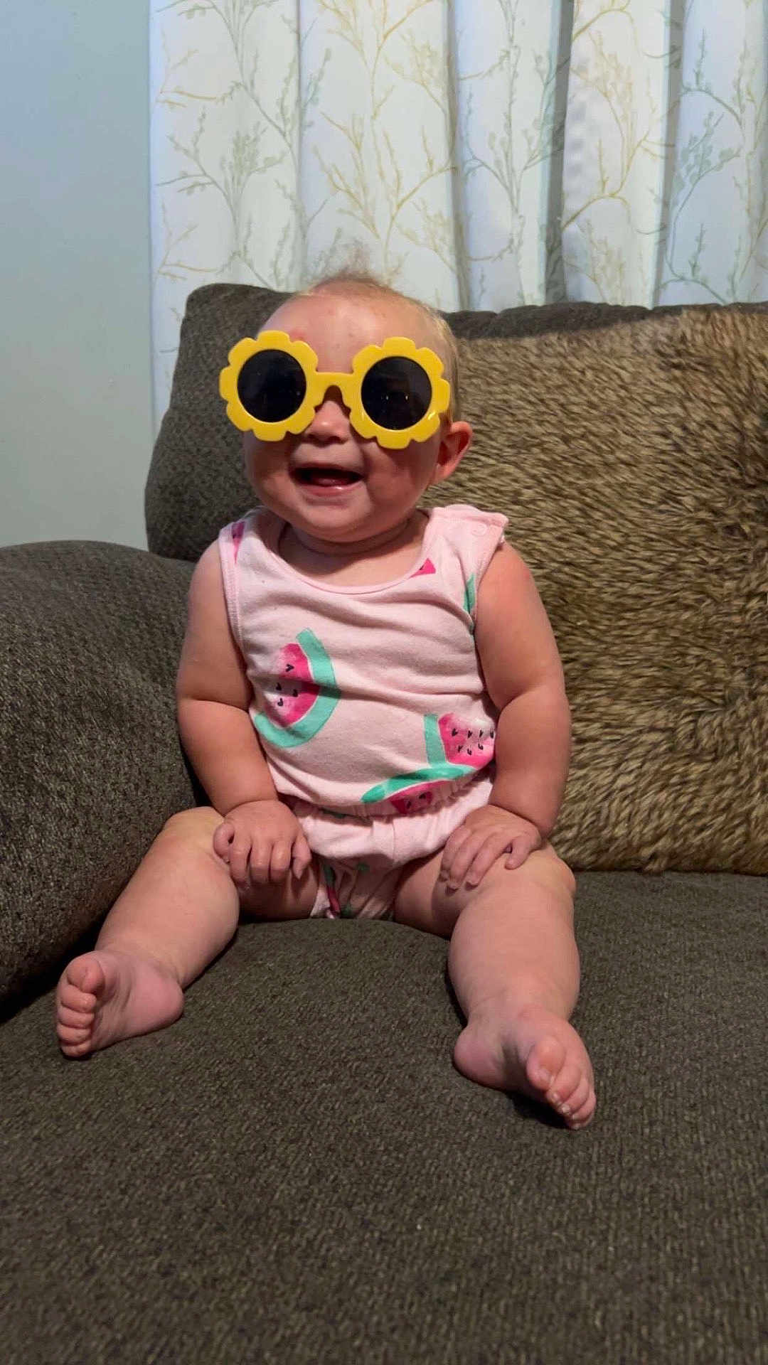 Lynnix is registered to the contest to win money with this photo: baby, child, sunglasses, yellow, pink_clothing, watermelon_pattern, smiling, sitting, couch, brown_couch, curtains, indoor, cute, happy, funny_glasses, toddler, face, legs, feet, home