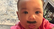 Zara is registered to the contest to win money with this photo: baby, child, pink_shirt, mustache, face, living_room, chair, dog, dog_crate, television, carpet, floor, wood_floor, furniture, indoor, person, portrait, blurred, close_up, home