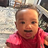 baby, child, pink_shirt, mustache, face, living_room, chair, dog, dog_crate, television, carpet, floor, wood_floor, furniture, indoor, person, portrait, blurred, close_up, home