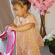 Harmonie a rejoint le concours — aidez-le/la à gagner de superbes lots ! toddler, child, pink_chair, floral_dress, flower_pot, pink_flowers, white_flowers, tile_floor, indoor, earrings, shoes, person, young_child, curly_hair, side_view, holding, standing, home_interior, plant, footwear
