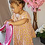 toddler, child, pink_chair, floral_dress, flower_pot, pink_flowers, white_flowers, tile_floor, indoor, earrings, shoes, person, young_child, curly_hair, side_view, holding, standing, home_interior, plant, footwear