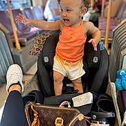 Ocianna joined the competition — help win amazing prizes! toddler, child, car_seat, orange_tank_top, white_shorts, curly_hair, bus_interior, handbag, water_bottle, person, leg, sneaker, seat, window, blurred_background, public_transport, happy, reaching, clothing, indoor