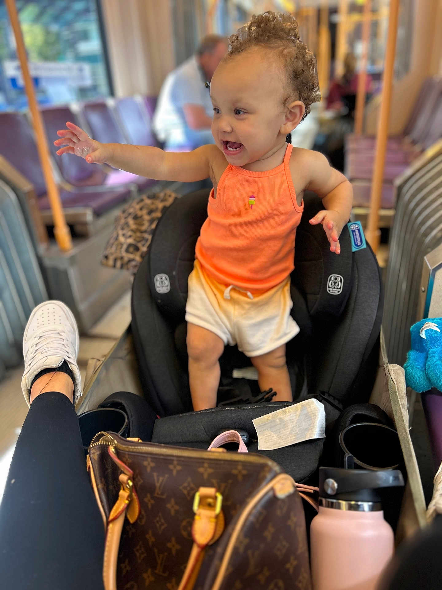 Ocianna joined the competition — help win amazing prizes! toddler, child, car_seat, orange_tank_top, white_shorts, curly_hair, bus_interior, handbag, water_bottle, person, leg, sneaker, seat, window, blurred_background, public_transport, happy, reaching, clothing, indoor