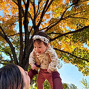 Alanna joined the competition — help win amazing prizes! baby, child, infant, adult, parent, outdoors, autumn, fall, trees, leaves, golden foliage, sky, family, lifting, smiling, headband, floral clothing, pants, portrait, sunlight