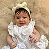 baby, child, infant, smiling, portrait, headband, bow, white_dress, eyelet_fabric, sitting, blanket, woven_texture, indoors, close_up, face, hands, brown_hair, happy, baby_fashion, cozy