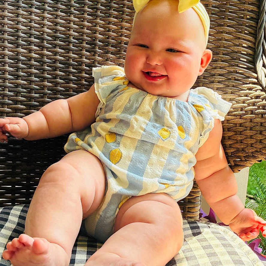 Cali joined the competition — help win amazing prizes! baby, child, smiling, yellow_bow, romper, striped_clothing, lemon_print, wicker_chair, checkered_cushion, outdoor, greenery, sunlight, cute, happy, infant, seated, plump_legs, headband, portrait, summer