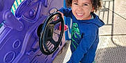 Daelyne is registered to the contest to win money with this photo: black_shoes, blue_hoodie, child, concrete_floor, curly_hair, daylight, dinosaur_print, fun, green_shorts, happy, imagination, metal_fence, outdoor, person, play, playground, purple_panel, smiling, steering_wheel, toy