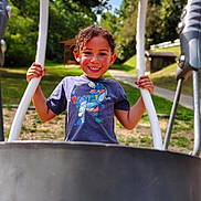 Daelyne is registered to the contest to win money with this photo: child, clouds, curly_hair, daytime, dinosaur_print, fun, greenery, happy, outdoor, park, person, play, playground, recreation, sky, smiling, sunlight, swing, trees, tshirt