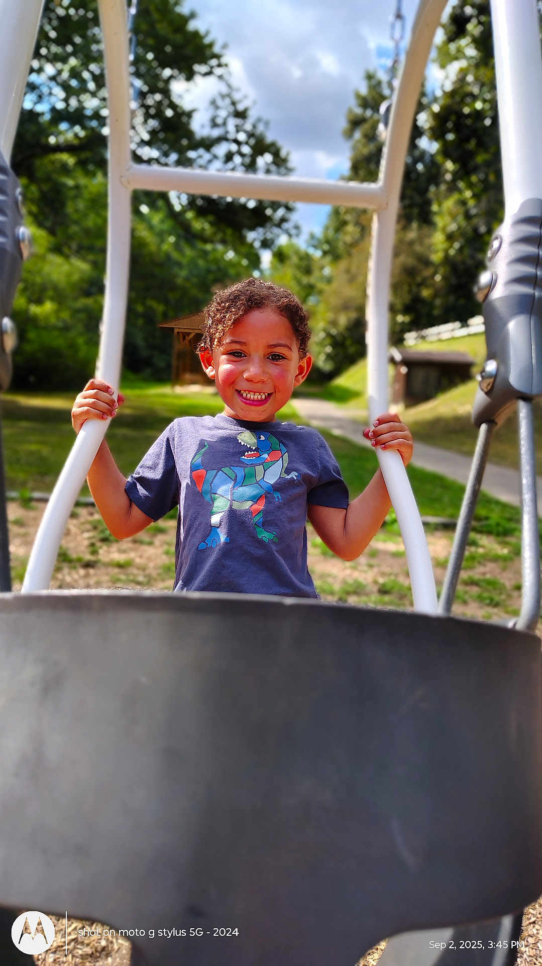 Daelyne is registered to the contest to win money with this photo: child, clouds, curly_hair, daytime, dinosaur_print, fun, greenery, happy, outdoor, park, person, play, playground, recreation, sky, smiling, sunlight, swing, trees, tshirt