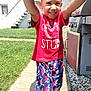 child, smiling, outdoor, grass, sidewalk, red_shirt, colorful_shorts, happy, person, grill, house, stairs, sunny, daylight, footwear, sock, patterned_clothing, reaching, playful, portrait