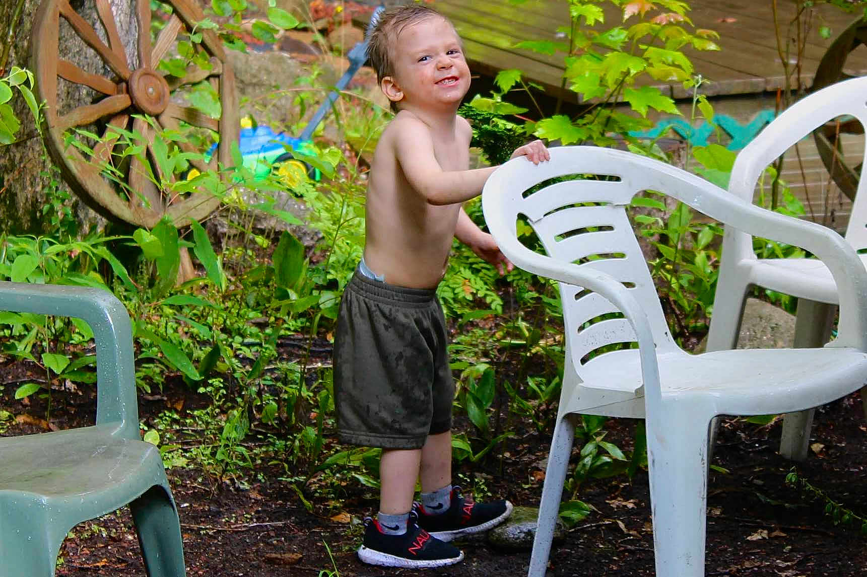 Jace is registered to the contest to win money with this photo: child, boy, outdoor, garden, plants, greenery, plastic_chair, sneakers, shorts, shirtless, smile, wet, dirt, tree, wagon_wheel, playful, nature, young, happy, person