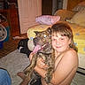 boy, dog, indoor, bed, pillow, carpet, fan, door, smile, red_eye, hugging, shirtless, floor, wood_paneling, furniture, blanket, pet, child, home, happy