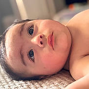 Antonio joined the competition — help win amazing prizes! baby, infant, face, eyes, eyelashes, nose, lips, cheek, skin, bare_chest, blanket, textured_surface, close_up, portrait, indoor, soft_lighting, bokeh, food_pouch, lying_down, curious