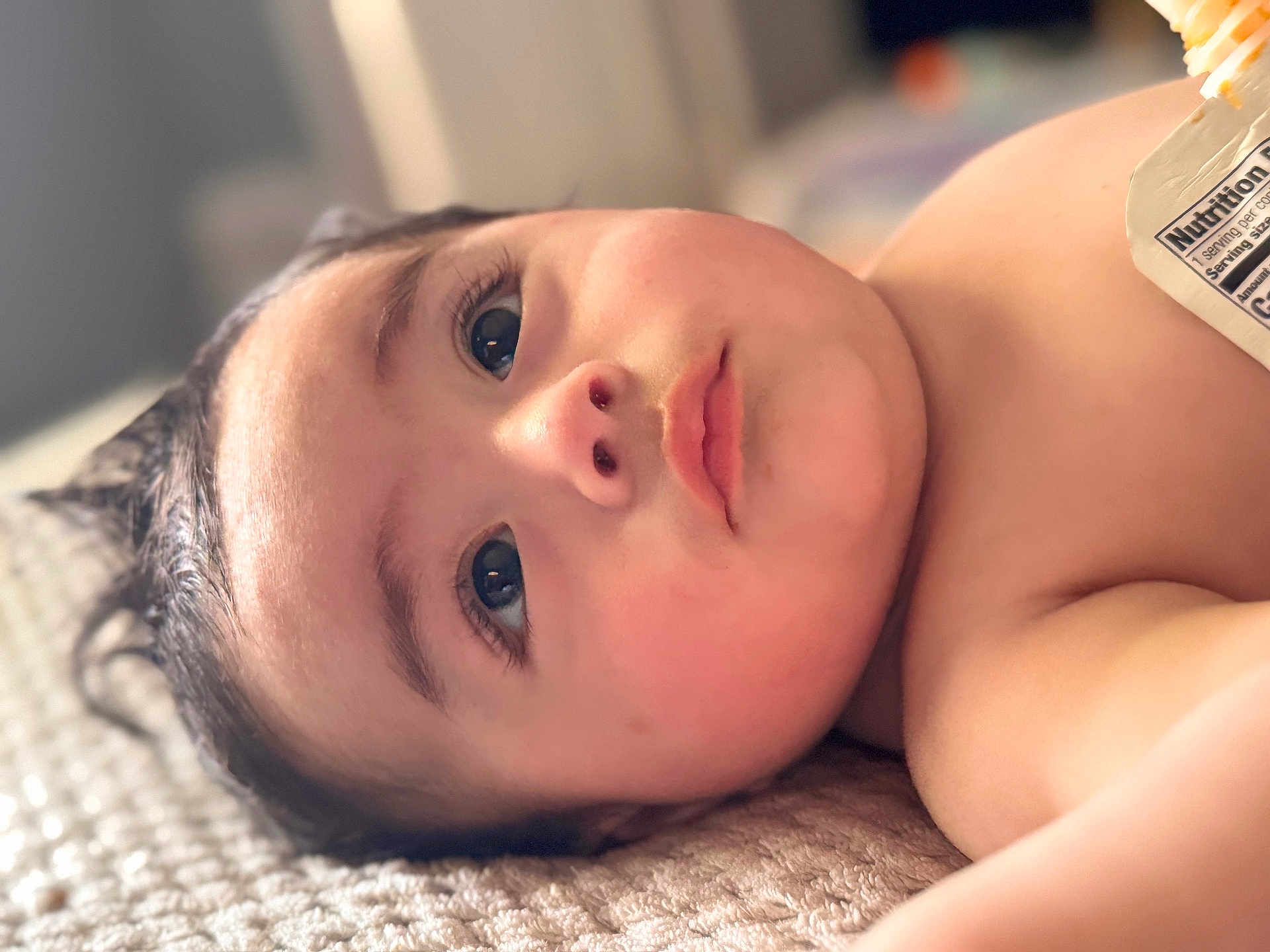 Antonio is registered to the contest to win money with this photo: baby, infant, face, eyes, eyelashes, nose, lips, cheek, skin, bare_chest, blanket, textured_surface, close_up, portrait, indoor, soft_lighting, bokeh, food_pouch, lying_down, curious
