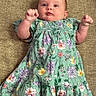Raelynn is registered to the contest to win money with this photo: baby, infant, child, dress, floral_pattern, green_dress, furniture, couch, blanket, skin, person, smiling, happy, indoors, portrait, cute, newborn, lying_down, fist, young