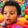 child, toddler, face, curly_hair, ball_pit, colorful_balls, play, expression, indoor, fun, young_child, portrait, bright_colors, plastic_balls, casual_clothing, closeup, happy, innocence, eye_contact, entertainment