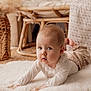 baby, infant, blue_eyes, lying_on_stomach, fluffy_rug, woven_mat, wicker_chair, neutral_colors, soft_texture, cozy, indoors, portrait, cute, chubby_cheeks, curious_expression, natural_light, nursery, white_clothing, hands, closeup