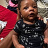 baby, infant, child, person, hand, clothing, onesie, black_clothing, text_on_clothing, indoor, bed, blanket, furniture, human_skin, face, eyes, mouth, expression, holding, room