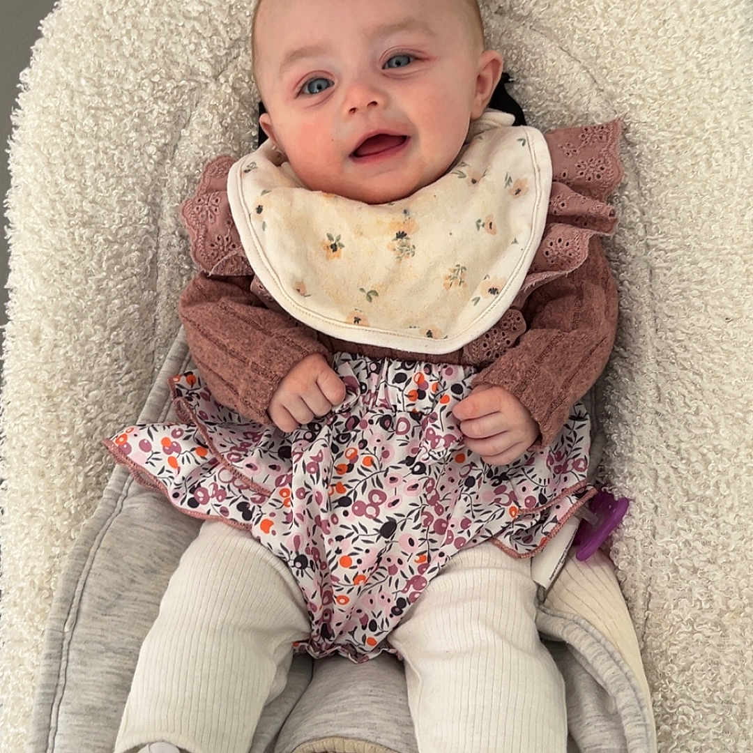 Inaya participe au concours pour gagner de l'argent avec cette photo : baby, infant, bib, sneakers, baby_seat, clothing, cute, smile, face, child, white_shoes, floral_pattern, warm_clothes, indoor, comfort, young_child, portrait, human, resting, cozy