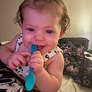 Blakely is registered to the contest to win money with this photo: baby, child, spoon, pink_bow, smiling, indoor, bed, blanket, floral_pattern, curly_hair, cute, holding, happy, portrait, person, clothing, skin, face, playful, infant