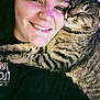 person, cat, tabby_cat, smiling, hugging, close_up, face, pet, animal, affection, cuddle, indoor, black_shirt, fur, whiskers, happy, relaxed, purple_light, portrait, companion