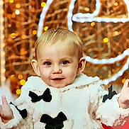 Aismée a rejoint le concours — aidez-le/la à gagner de superbes lots ! toddler, child, smiling, face, coat, white, black_bows, hands, indoors, portrait, cute, happy, baby, warm_lighting, decorative_lights, fashion, winter_clothing, expression, young_child, festive