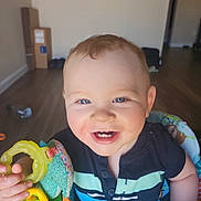 Dante joined the competition — help win amazing prizes! baby, child, smiling, blue_eyes, toy, baby_seat, indoor, floor, wooden_floor, clothing, short_sleeves, person, happy, cute, infant, hand, face, home, background, teeth