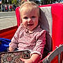 toddler, child, smiling, stroller, red, phone, floral_pattern, outdoor, daylight, face, hair, clothing, hand, park, flag, people, tree, happy, person, cute