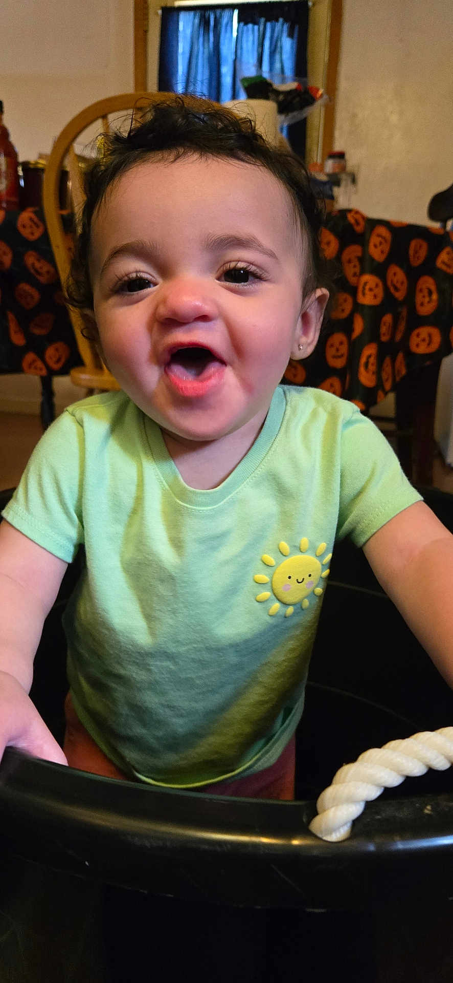 Delilah joined the competition — help win amazing prizes! toddler, child, curly_hair, green_shirt, sun_patch, smiling, indoor, table, pumpkin_decor, black_container, rope_handle, happy, face, person, clothing, chair, background, home, cute, expression