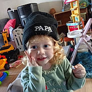 émeraude a rejoint le concours — aidez-le/la à gagner de superbes lots ! toddler, child, beanie, hat, curly_hair, indoor, toy, playroom, smiling, green_shirt, person, furniture, floor, wooden_floor, plastic_toys, chair, couch, carpet, happy, cute