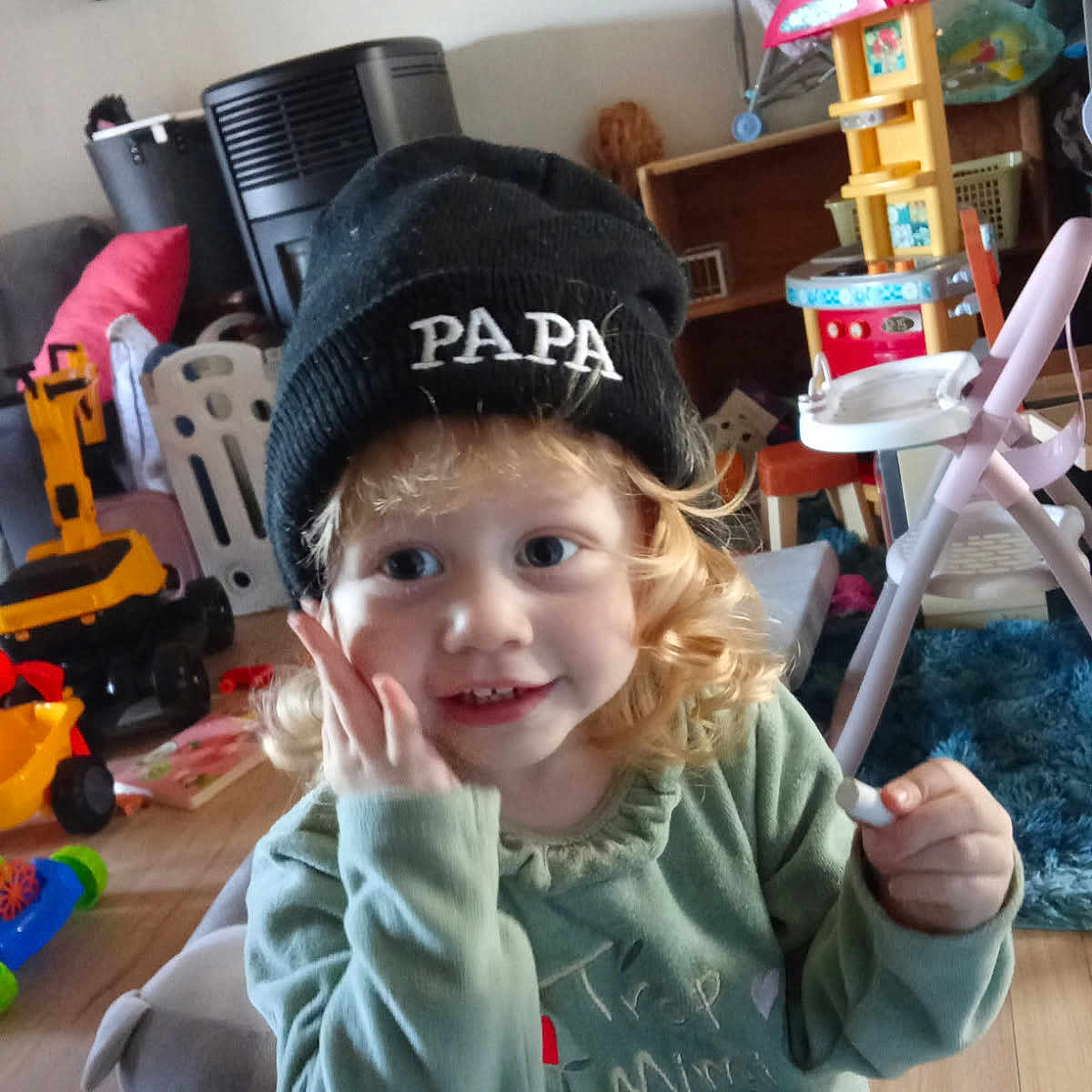 émeraude a rejoint le concours — aidez-le/la à gagner de superbes lots ! beanie, carpet, chair, child, couch, curly_hair, cute, floor, furniture, green_shirt, happy, hat, indoor, person, plastic_toys, playroom, smiling, toddler, toy, wooden_floor