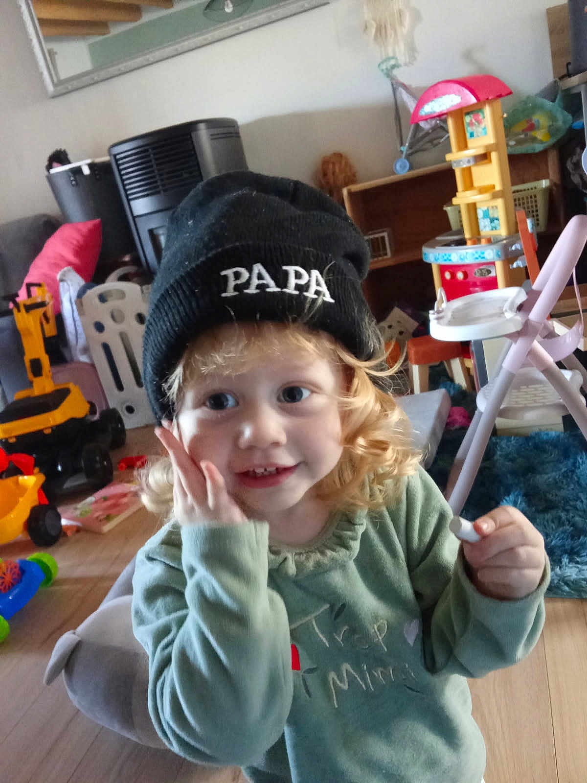 émeraude a rejoint le concours — aidez-le/la à gagner de superbes lots ! toddler, child, beanie, hat, curly_hair, indoor, toy, playroom, smiling, green_shirt, person, furniture, floor, wooden_floor, plastic_toys, chair, couch, carpet, happy, cute