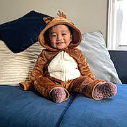 Sebastian is registered to the contest to win money with this photo: toddler, child, reindeer_costume, onesie, smiling, indoors, couch, blue_couch, pillows, brown_clothing, white_pouch, happy, cute, cozy, window, socks, plush, young_child, home, portrait
