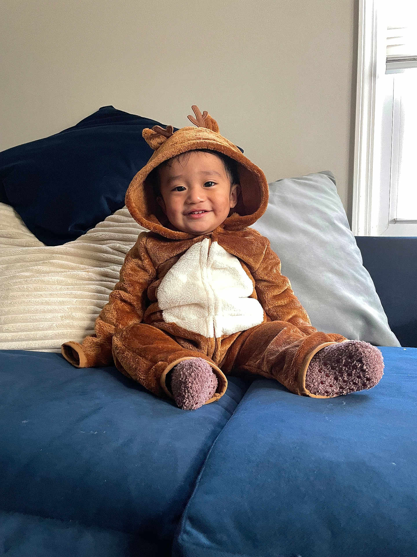 Sebastian is registered to the contest to win money with this photo: toddler, child, reindeer_costume, onesie, smiling, indoors, couch, blue_couch, pillows, brown_clothing, white_pouch, happy, cute, cozy, window, socks, plush, young_child, home, portrait