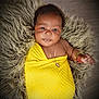 adorable, baby, bracelet, child, cozy, cute, face, fluffy_rug, gold_jewelry, hand, indoor, infant, lying_down, necklace, portrait, skin, smiling, soft_texture, wrapped, yellow_blanket