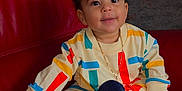 Liam a rejoint le concours — aidez-le/la à gagner de superbes lots ! toddler, child, smiling, sitting, colorful_clothing, sneakers, red_couch, indoor, gold_chain, curly_hair, happy, young_child, portrait, fashion, cute, leather_furniture, playful, casual_wear, person, home