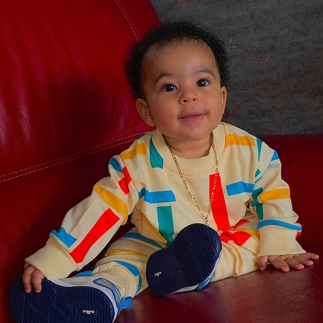 Liam a rejoint le concours — aidez-le/la à gagner de superbes lots ! casual_wear, child, colorful_clothing, curly_hair, cute, fashion, gold_chain, happy, home, indoor, leather_furniture, person, playful, portrait, red_couch, sitting, smiling, sneakers, toddler, young_child