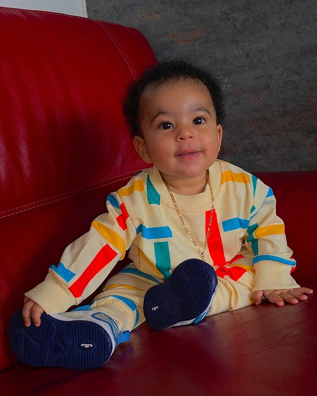 Liam a rejoint le concours — aidez-le/la à gagner de superbes lots ! toddler, child, smiling, sitting, colorful_clothing, sneakers, red_couch, indoor, gold_chain, curly_hair, happy, young_child, portrait, fashion, cute, leather_furniture, playful, casual_wear, person, home
