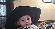 Skyler joined the competition — help win amazing prizes! baby, child, cowboy_hat, onesie, person, hands, indoor, couch, wall, painting, shelf, welcome_sign, curtain, window, orange_shirt, patterned_clothing, holding, face, infant, cozy_room