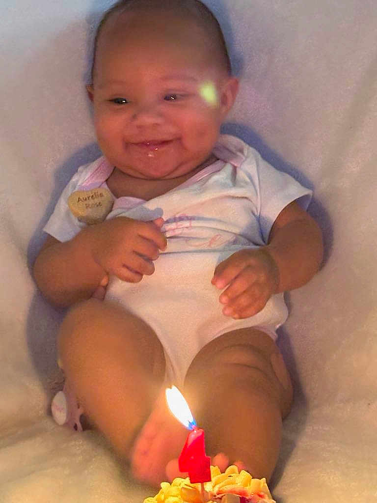 Aurelia is registered to the contest to win money with this photo: baby, child, smiling, birthday_candle, candle_flame, cupcake, dimples, white_clothing, sitting, soft_surface, celebration, closeup, cute, infant, indoors, happy, small_hands, legs, face, warm_light