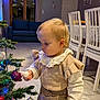 toddler, child, christmas_tree, ornament, holiday, indoor, curious, decorations, plaid_skirt, ruffled_sleeves, cozy, home, furniture, dining_chairs, lights, festive, blonde_hair, floor, curtains, sofa