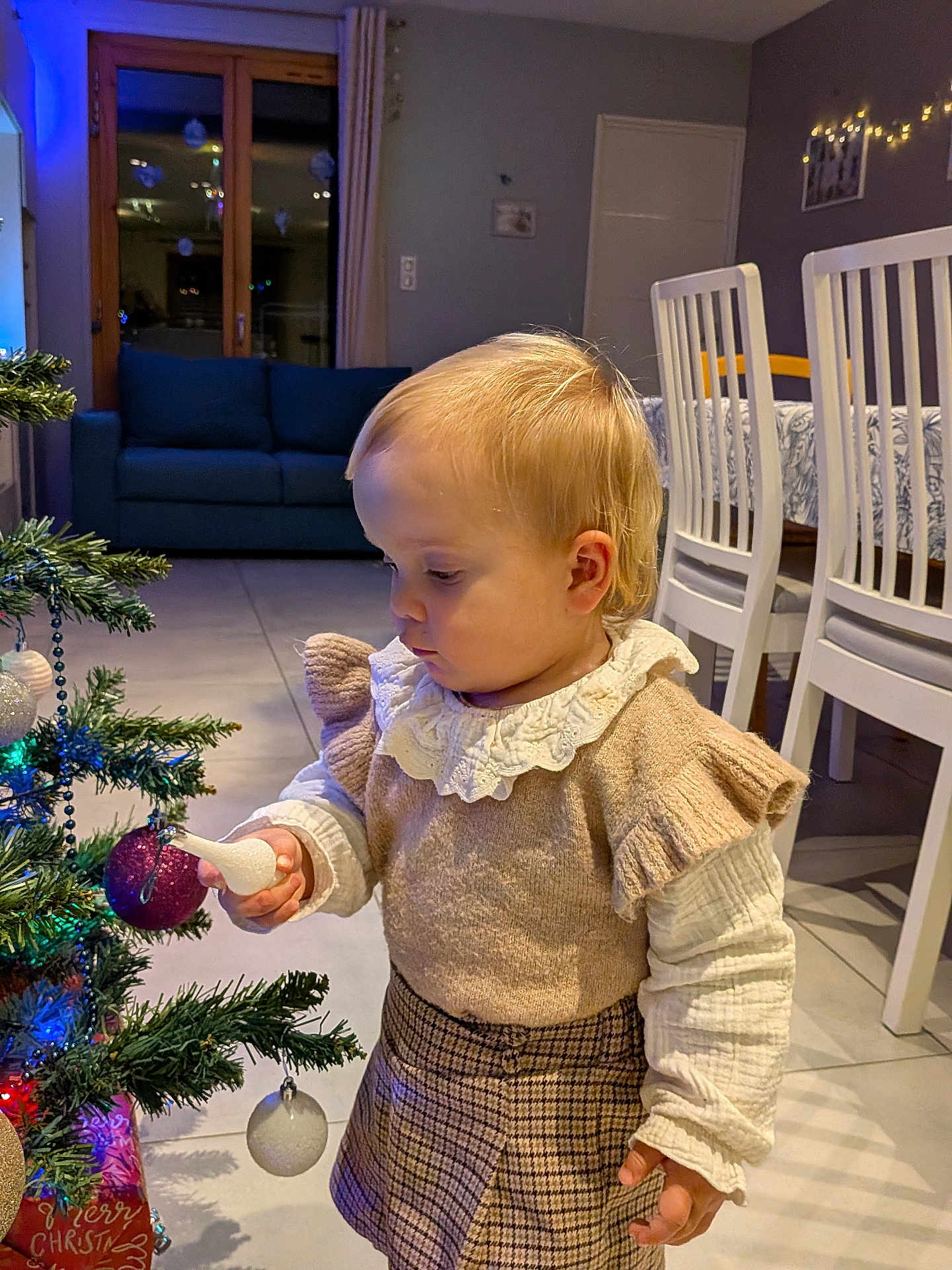 Sélénia participe au concours pour gagner de l'argent avec cette photo : toddler, child, christmas_tree, ornament, holiday, indoor, curious, decorations, plaid_skirt, ruffled_sleeves, cozy, home, furniture, dining_chairs, lights, festive, blonde_hair, floor, curtains, sofa