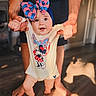 Hazel joined the competition — help win amazing prizes! baby, child, infant, person, hands, floor, wood_floor, bow, headwear, clothing, onesie, cartoon_character, minnie_mouse, smiling, walking, indoor, adult, supporting_hands, feet, natural_light