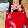 baby, child, pacifier, toy_car, indoor, floor_tiles, pink_clothing, seat, steering_wheel, face, eyes, hair, car_toy, smile_painting, playtime, infant, cute, expression, seat_belt, colorful