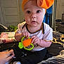 Oaklynn joined the competition — help win amazing prizes! baby, child, toy, orange_bow, carrot_toy, indoor, bed, blanket, clothing, sitting, hand, person, cute, infant, white_shirt, black_pants, sock, home, curious, wide_eyes