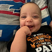Oyla is registered to the contest to win money with this photo: baby, child, infant, smiling, happy, portrait, face, hand, fist, bed, bedspread, blanket, indoor, close_up, black_shirt, text_on_clothing, casual, lying_down, playful, photography