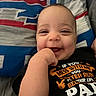 baby, child, infant, smiling, happy, portrait, face, hand, fist, bed, bedspread, blanket, indoor, close_up, black_shirt, text_on_clothing, casual, lying_down, playful, photography