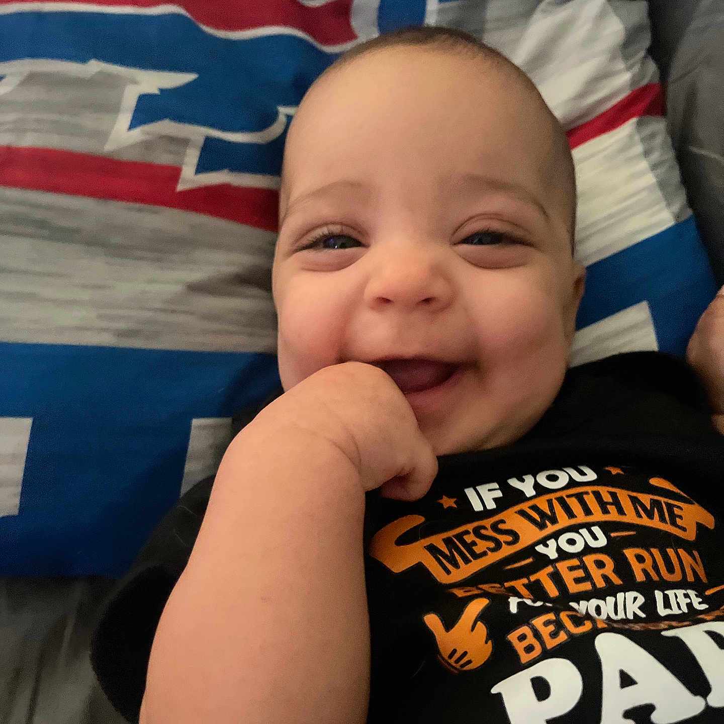 Oyla is registered to the contest to win money with this photo: baby, child, infant, smiling, happy, portrait, face, hand, fist, bed, bedspread, blanket, indoor, close_up, black_shirt, text_on_clothing, casual, lying_down, playful, photography