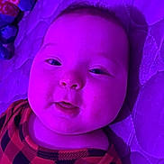 Malachi is registered to the contest to win money with this photo: baby, child, infant, portrait, face, eyes, smiling, lying_down, blanket, bed, clothing, checkered_pattern, purple_lighting, indoor, close_up, adorable, head, person, photography, relaxed