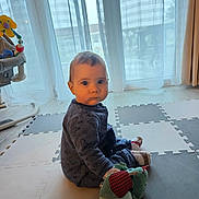 Maël a rejoint le concours — aidez-le/la à gagner de superbes lots ! baby, child, indoor, toy, stuffed_animal, foam_mat, sitting, curtains, floor, clothing, person, young_child, play_area, sock, head, face, eyes, furniture, home, light